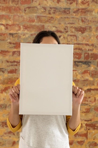 front view close up of a young caucasian woman holding a blank white canvas in her hands awhich covers her face, standing in front of a brick wall