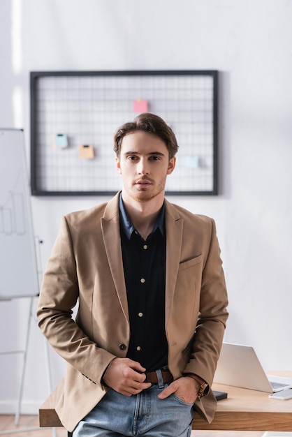 Front view of confident businessman with hand in pocket looking at camera while leaning on desk on