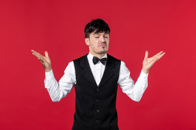 Front view of confused male waiter in a uniform with butterfly on neck and questioning about something on red wall