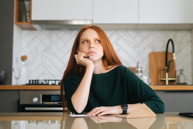 Front view of confused young woman writing handwritten letter sitting at table with envelope in