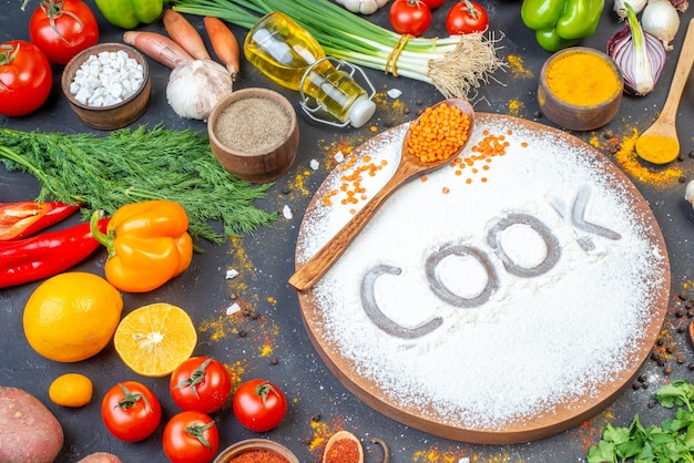 Front view of cook writing with flour on wooden board among fresh vegetables spices green bundles fallen oil bottle on black wall