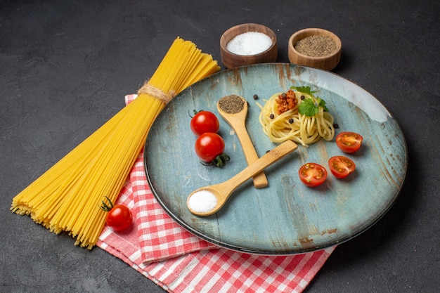 Вид спереди вкусных спагетти, подаваемых с мясной зеленью, помидорами, солью, перцем на тарелке и сырой пастой на красном полосатом полотенце на черном фоне