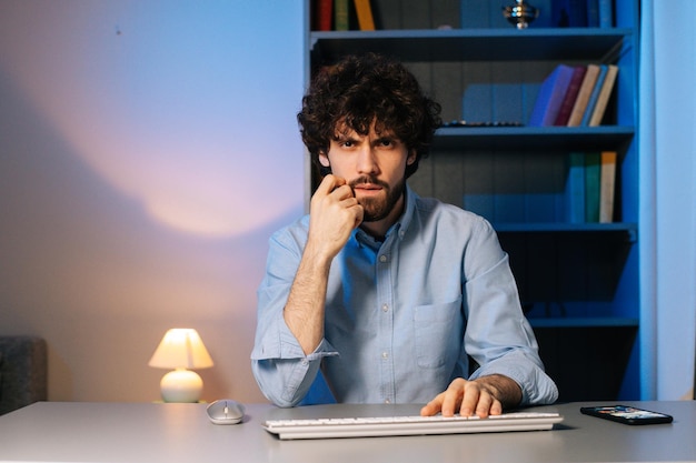 Front view of disgruntled young man typing online message on wireless keyboard looking at camera