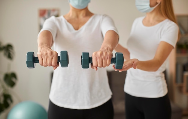 Front view of elder woman with medical mask in covid recovery doing exercises with dumbbells