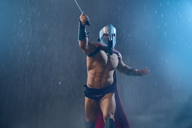 Front view of fearless wet roman gladiator in iron helmet attacking with sword. Muscular screaming shirtless spartan in red cloak and armor running during fight in rainy bad weather.