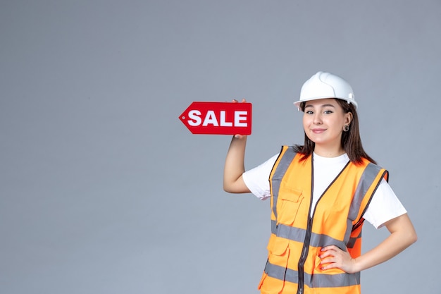 Front view of female builder holding red sale board on gray wall