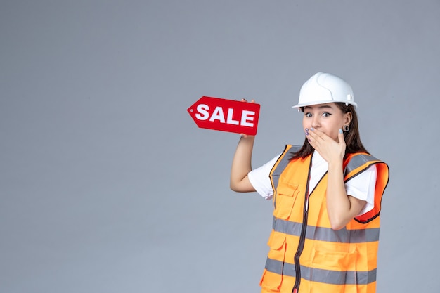 Front view of female builder holding red sale board on gray wall