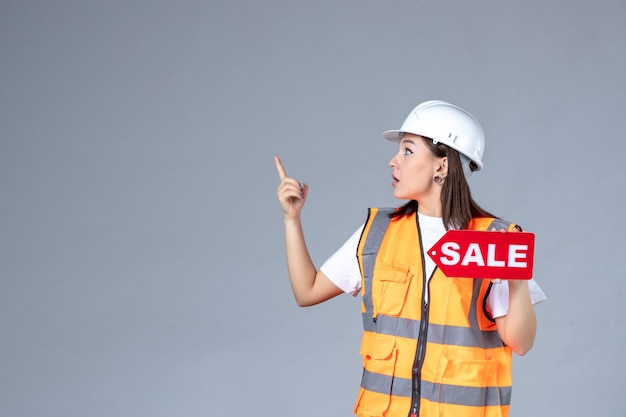 Front view of female builder holding red sale board on gray wall