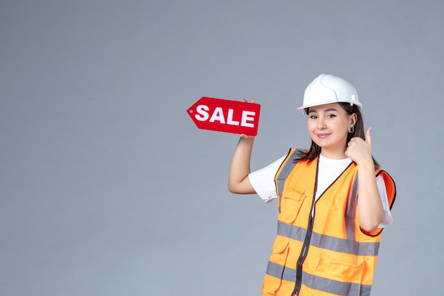 Front view of female builder holding red sale board on gray wall