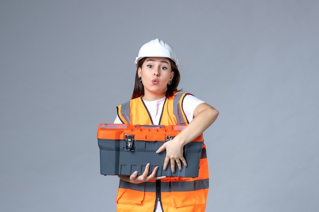 Front view of female builder holding tool case on gray wall