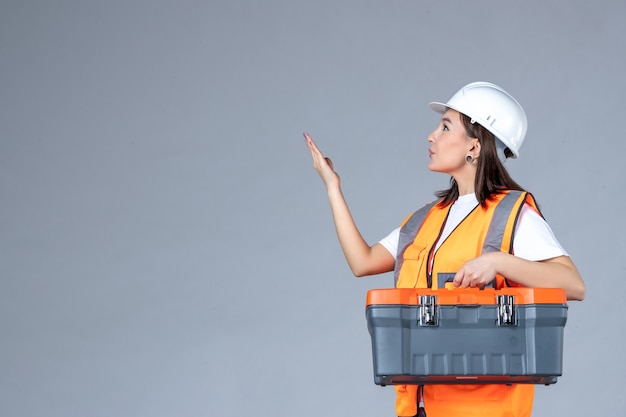 Front view of female builder holding tool case on gray wall