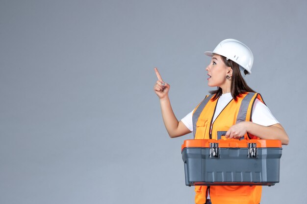 Front view of female builder holding tool case on gray wall