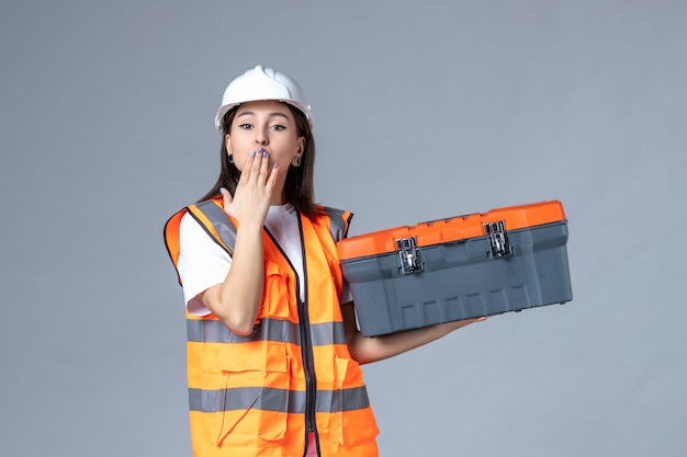 Front view of female builder holding tool case on gray wall
