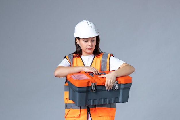 Front view of female builder holding tool case on gray wall