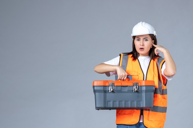 Front view of female builder with heavy tool case on gray wall