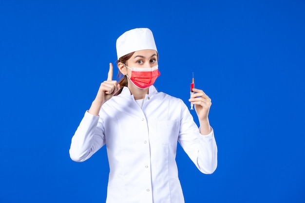 Front view female nurse in white medical suit with red mask and injection in her hands on blue 