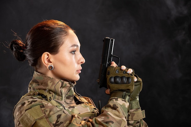 Vista frontale della soldatessa con la pistola in uniforme sul muro nero