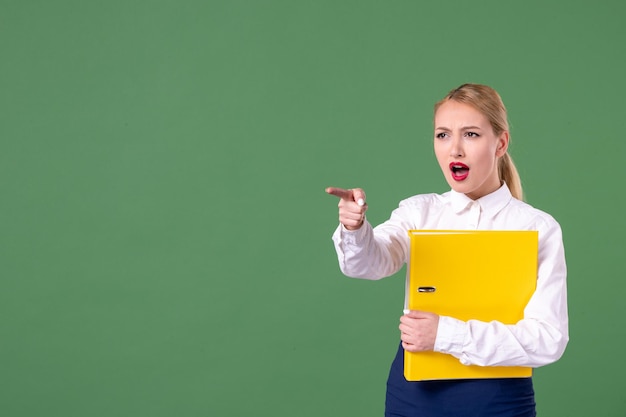 Front view female teacher holding yellow files and pointing on green background lesson book study library school university uniform students