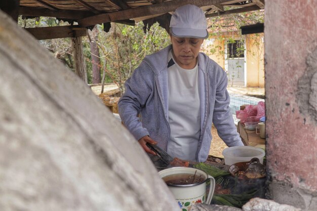 Front view of a fifty year old adult woman cooking meat and\
vegetables on a wood stove under a wooden kiosk with trees and a\
yellow house behind