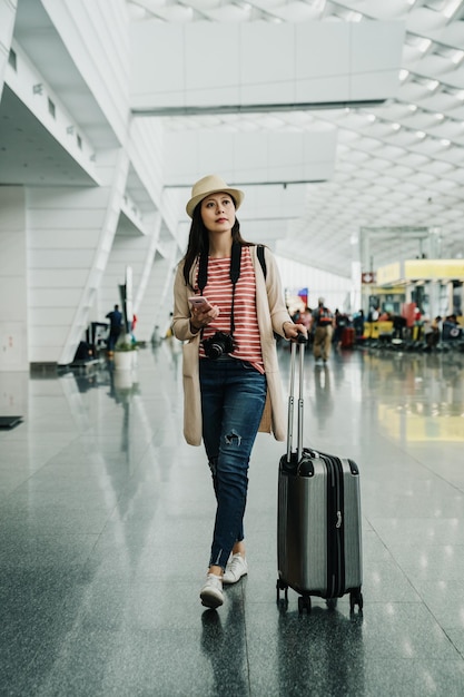 Front view of full length traveler woman walking and using\
smart phone in airport lobby terminal. young girl in hat carrying\
camera and luggage finding direction of flight gate while\
departure.