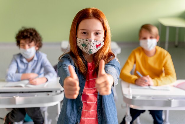 Foto vista frontale della ragazza con mascherina medica in aula che mostra i pollici in su