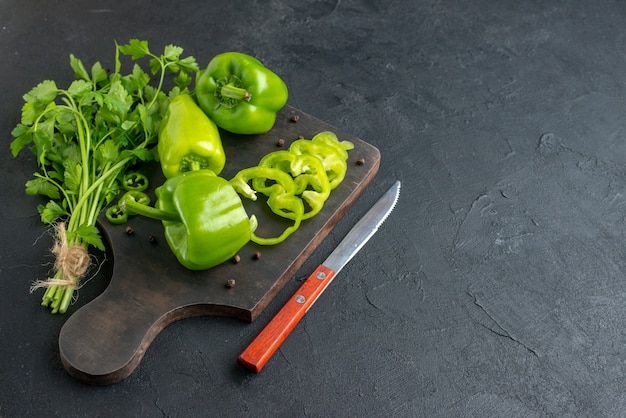 Front view of green bundle fresh whole green peppers on wooden cutting board knife on black distressed surface