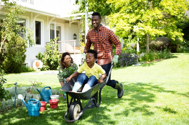 Front view of a happy African American couple and their son in the garden, the man pushing his son in a wheelbarrow while the mother watches. Family enjoying time at home, lifestyle concept