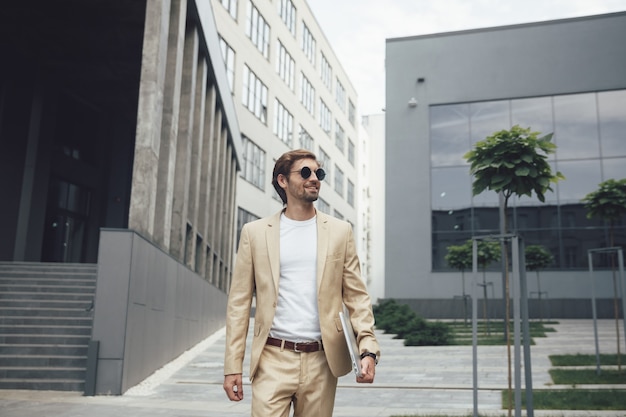 Front view of happy bearded businessman walking with personal laptop on street