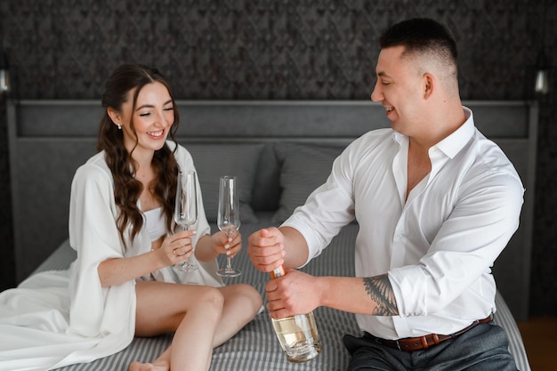 Front view of happy brides couple sitting in apartment on bed woman dressed in white silk robe holding glasses while men opening champagne Bride with groom celebrating wedding day