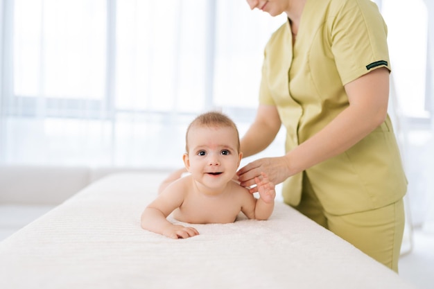 Front view of happy infant baby boy looking at camera lying on belly while unrecognizable female masseuse performing professional massage for back Loving mom hands doing back massage to newborn son