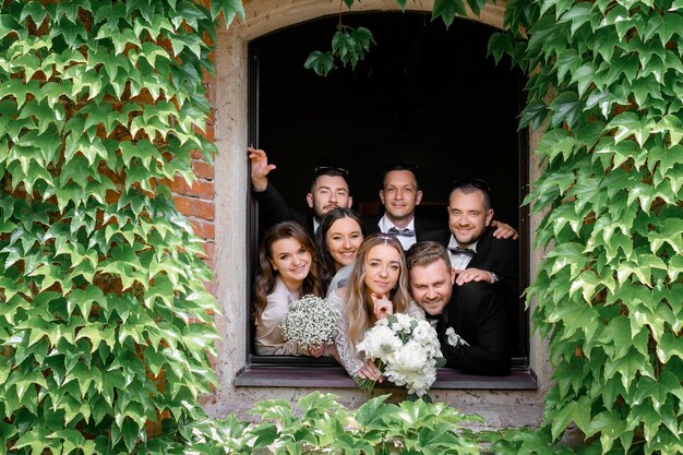 Vista frontale di una coppia sposata felice con ragazze e ragazzi in piedi e in posa davanti alla telecamera dalla finestra che si trova in un edificio antico coperto da piantexa