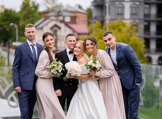 Front view of happy newlyweds and their friends wearing in stylish suits and gowns holding bouquets of flowers hugging each