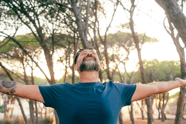 Front view of happy overjoyed mature man outstretching arms and enjoying outdoor leisure activity with park trees in background People and happiness Joyful male opening arms hugging life Joyful