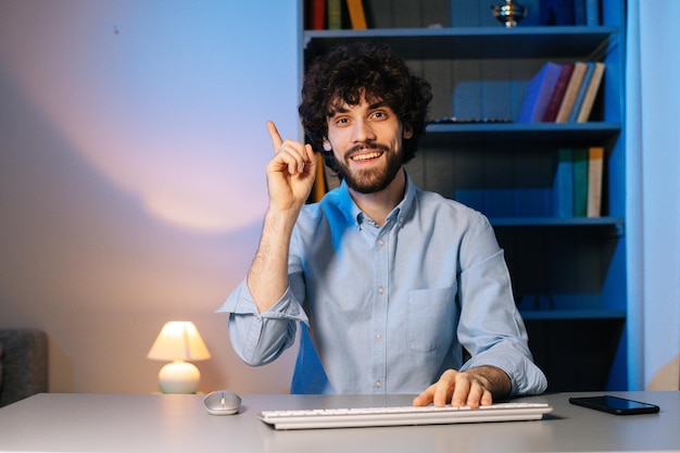 Front view of happy young man having idea moment pointing
finger up while sitting at desk with computer at home office.
smiling happy bearded guy showing eureka gesture. concept of remote
working.