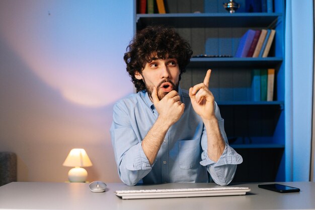 Front view of happy young man having idea moment pointing finger up while sitting at desk with computer at home office. Smiling happy bearded guy showing eureka gesture. Concept of remote working.