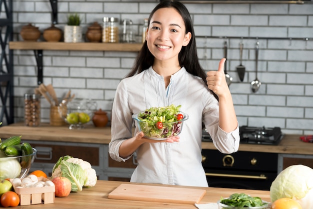 Photo front view of happy young woman holding glass of bowl with salad showing thumb up sign in kitchen