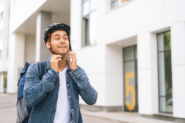 Front view of joyful young courier male with thermo backpack putting on helmet on head before to deliver an order to customer by bicycle. Handsome delivery man begins delivery of order to client