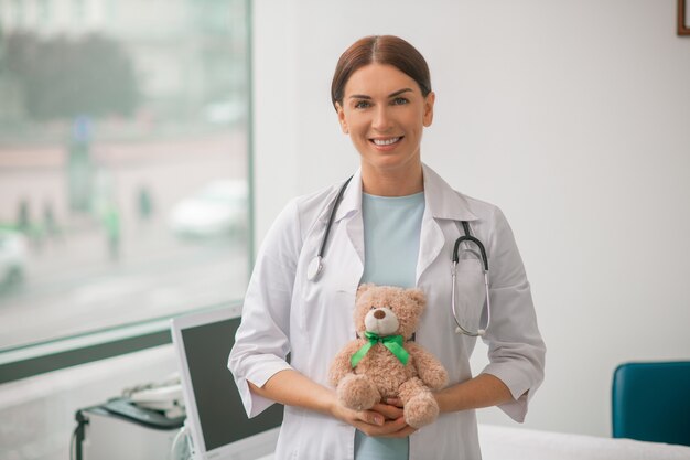 Front view of a joyous female doctor with a soft toy and a stethoscope standing at her office