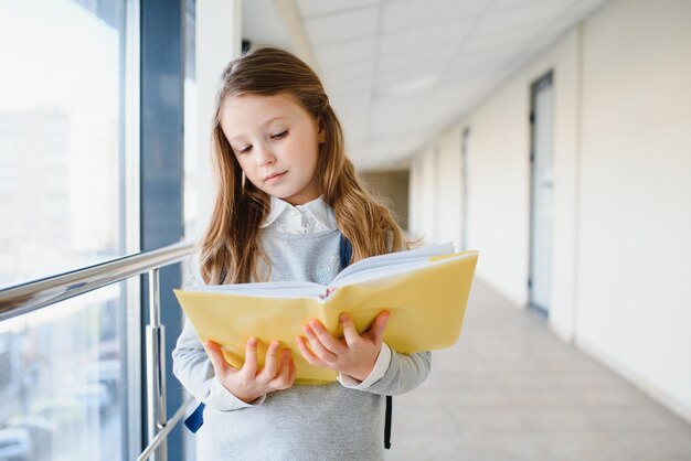 Photo front view of little beautiful school girl among corridor at school holding notes at hands funny and happy girl smiling at camera resting after lessons on primary school