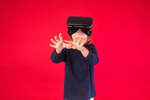 Front view of little girl wearing virtual reality glasses amazed and touching with her hands virtually on red background Virtual augmented reality technology games and childhood concept