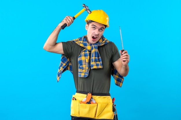 front view male builder in helmet holding screwdriver and hammer on blue background flat house job buildings worker constructor