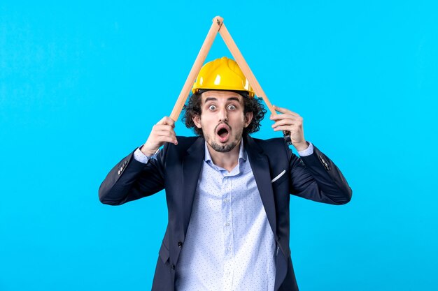 Front view male builder in helmet holding wooden geometric\
figure on blue background work building job business architecture\
constructor