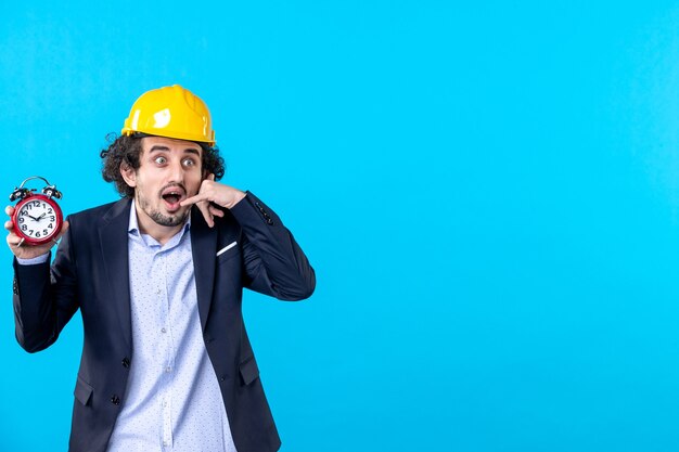 Front view male builder in helmet and suit holding clocks on blue background business design work architecture constructor buildings time job