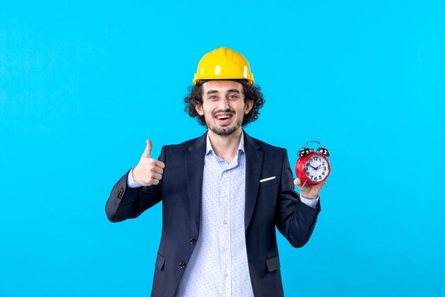 front view male builder in helmet and suit holding clocks on blue background constructor building architecture work job design