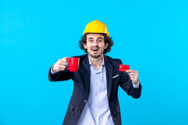 front view male builder in suit holding bank card and cup on blue background business building work constructor tea job money