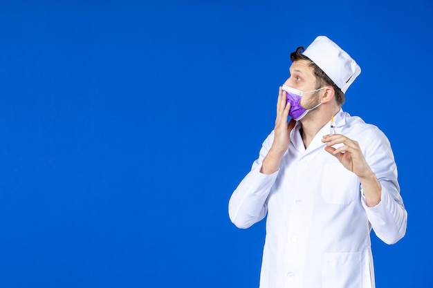 Front view of male doctor in medical suit and mask holding injection on blue 