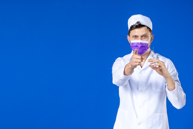 Vista frontale del medico maschio in tuta medica e maschera che tiene vaccino e iniezione sull'azzurro