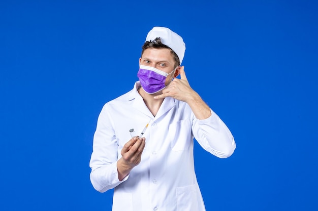 Front view of male doctor in medical suit and mask with injection and vaccine on blue 