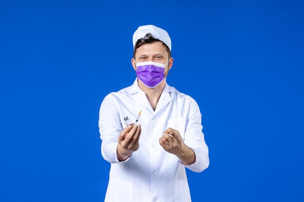 Front view of male doctor in medical suit and mask with injection and vaccine on blue 