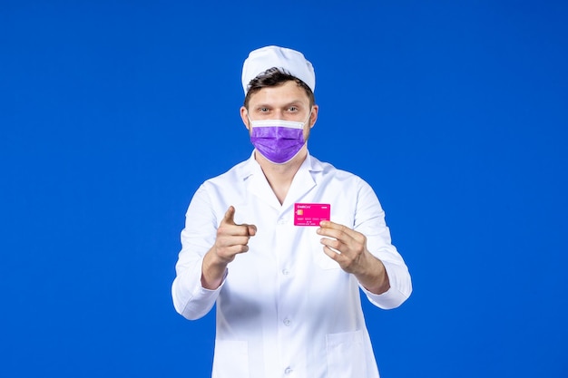 Front view of male doctor in medical suit and purple mask holding credit card on blue 